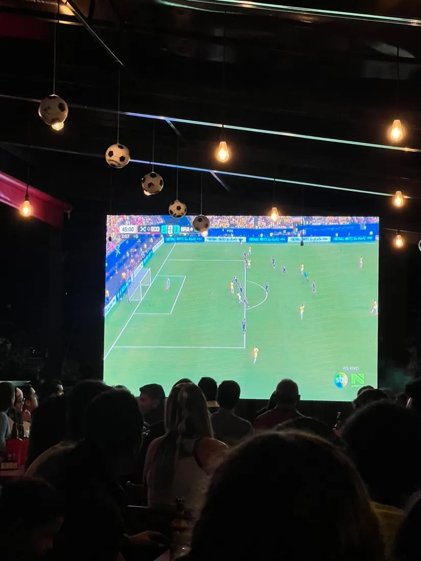 Jogo de futebol passando no telão do Josepio, com bolas de futebol penduradas no teto e a casa cheia